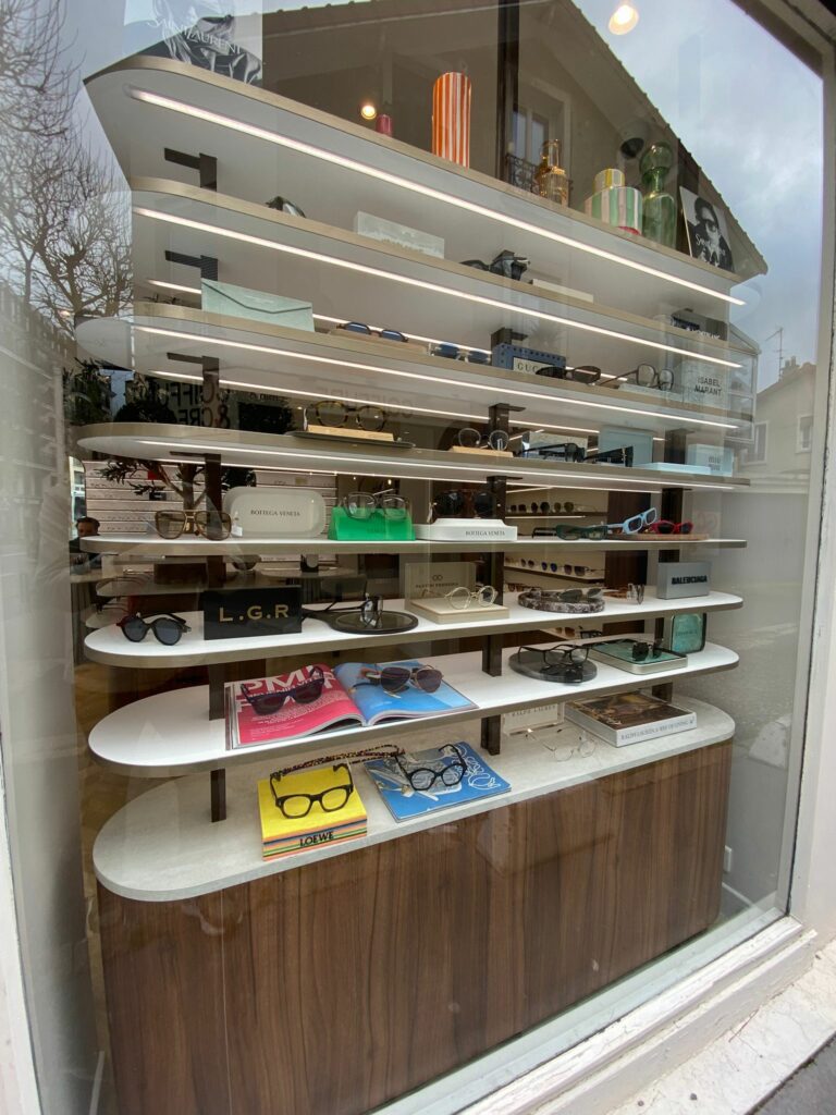 Vitrine de magasin d’optique avec étagères design rétroéclairées et mise en scène de lunettes de marques dans un mobilier haut de gamme.