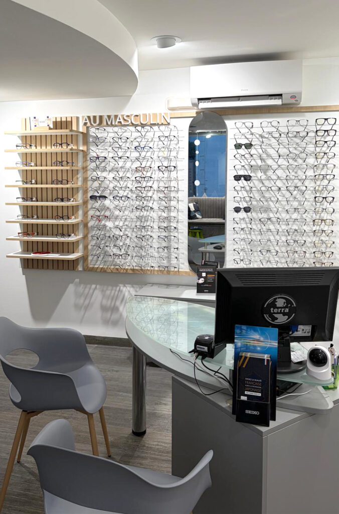 Espace conseil optique avec bureau, assises clients et mur de présentation de lunettes “au masculin” avec miroir intégré et éclairage soigné.