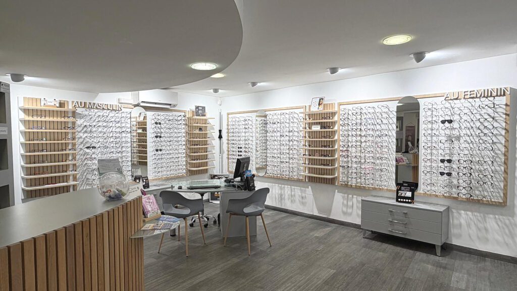 Agencement de magasin d’optique avec murs de présentation de lunettes “au masculin” et “au féminin”, espace de vente central et mobilier bois contemporain.
