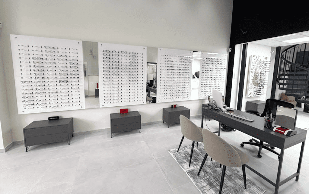 Agencement de magasin d’optique avec mur de présentation de lunettes, miroirs intégrés et espace de conseil avec bureau et assises modernes.