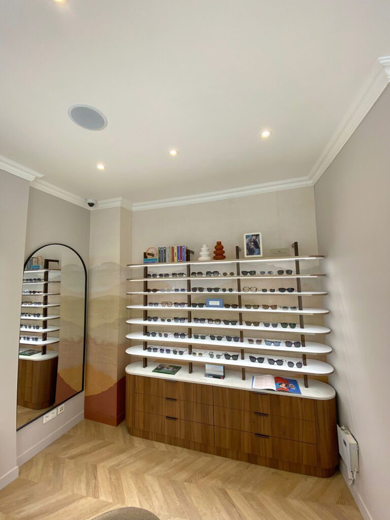 Agencement de magasin d’optique avec présentoir mural de lunettes, étagères rétroéclairées et grand miroir dans un espace chaleureux en bois.