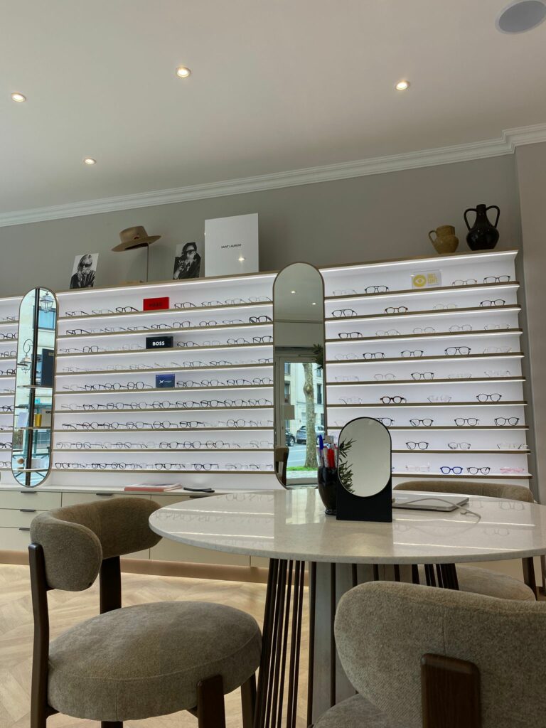 Magasin d’optique contemporain avec mur de présentation de lunettes rétroéclairé et table centrale de conseil client.