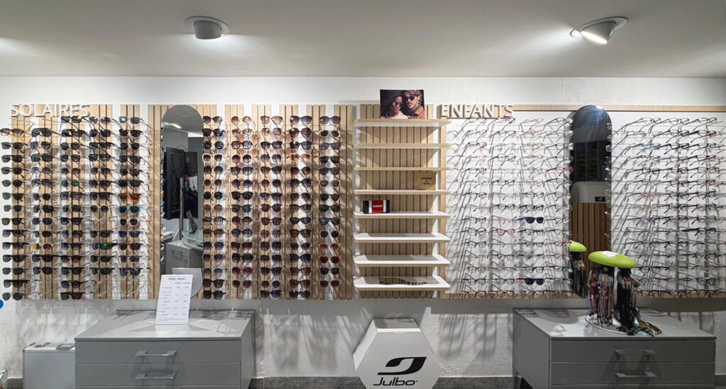 Présentoir mural de lunettes solaires et espace enfants dans un magasin d’optique avec mobilier bois, rangements et organisation par univers.