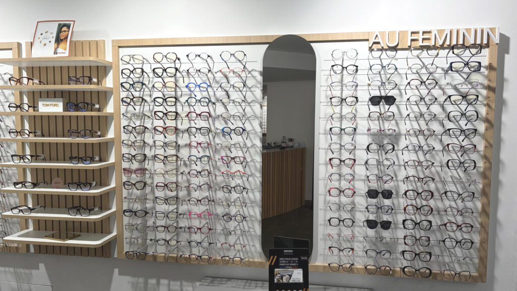 Mur de présentation de lunettes “au féminin” avec miroir central, mobilier bois et mise en valeur des montures optiques.