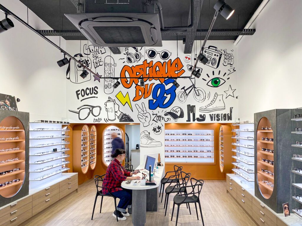 Magasin d’optique design avec fresque murale graphique, présentoirs de lunettes rétroéclairés et espace central de conseil client.