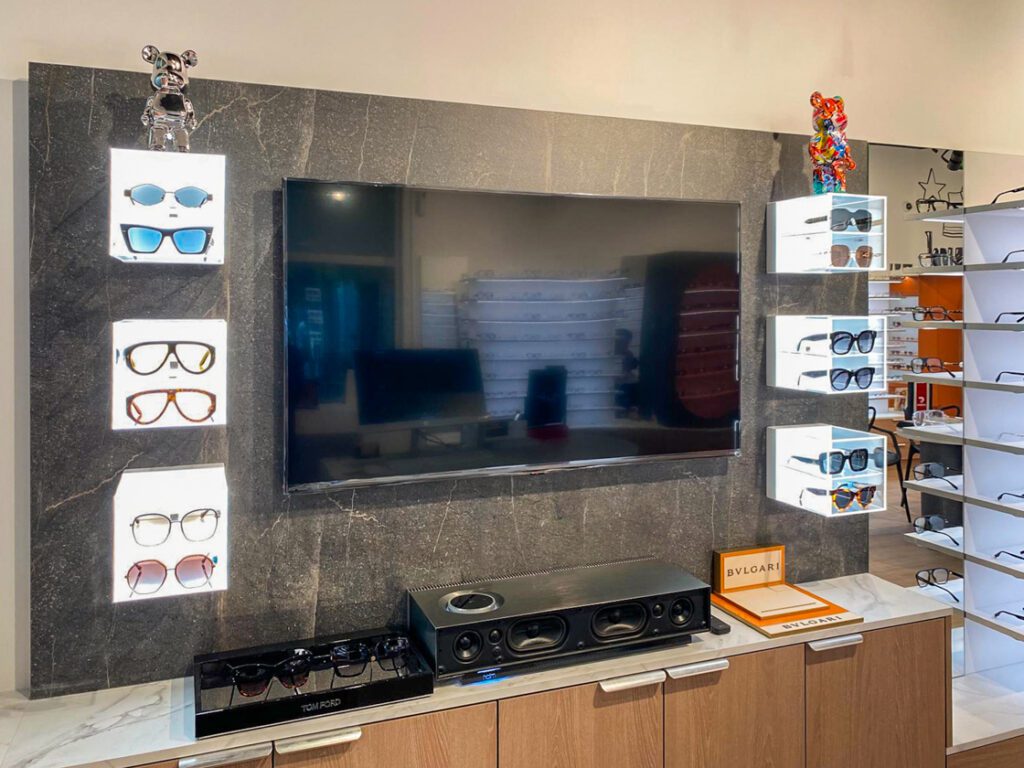Mobilier d’agencement optique avec mur décoratif, écran intégré et niches lumineuses mettant en valeur des lunettes et accessoires.