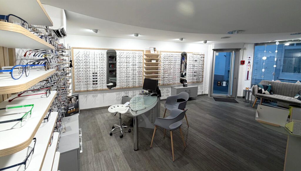 Vue intérieure d’un magasin d’optique avec comptoir central, murs de lunettes et espace d’accueil proche de l’entrée vitrée.