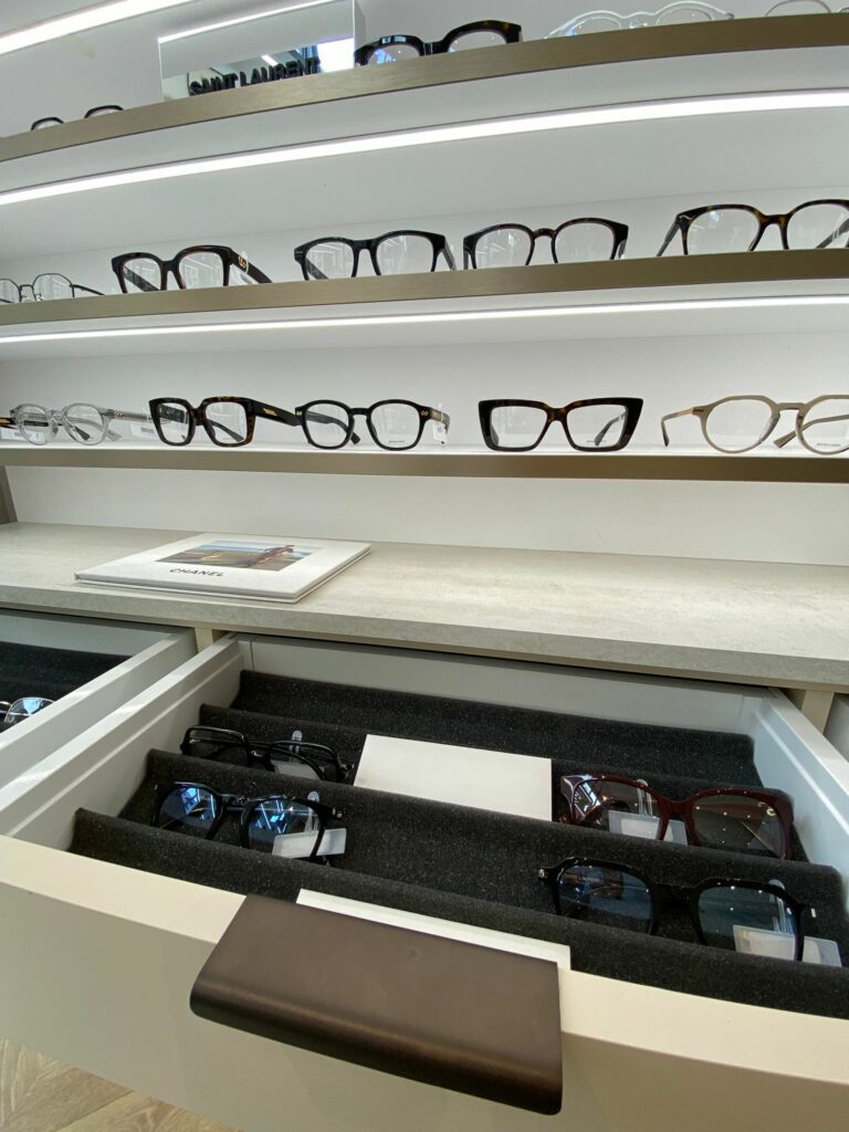 Entrée de magasin d’optique avec présentoir central, arbre décoratif et mobilier bois sur parquet pour une ambiance chaleureuse.